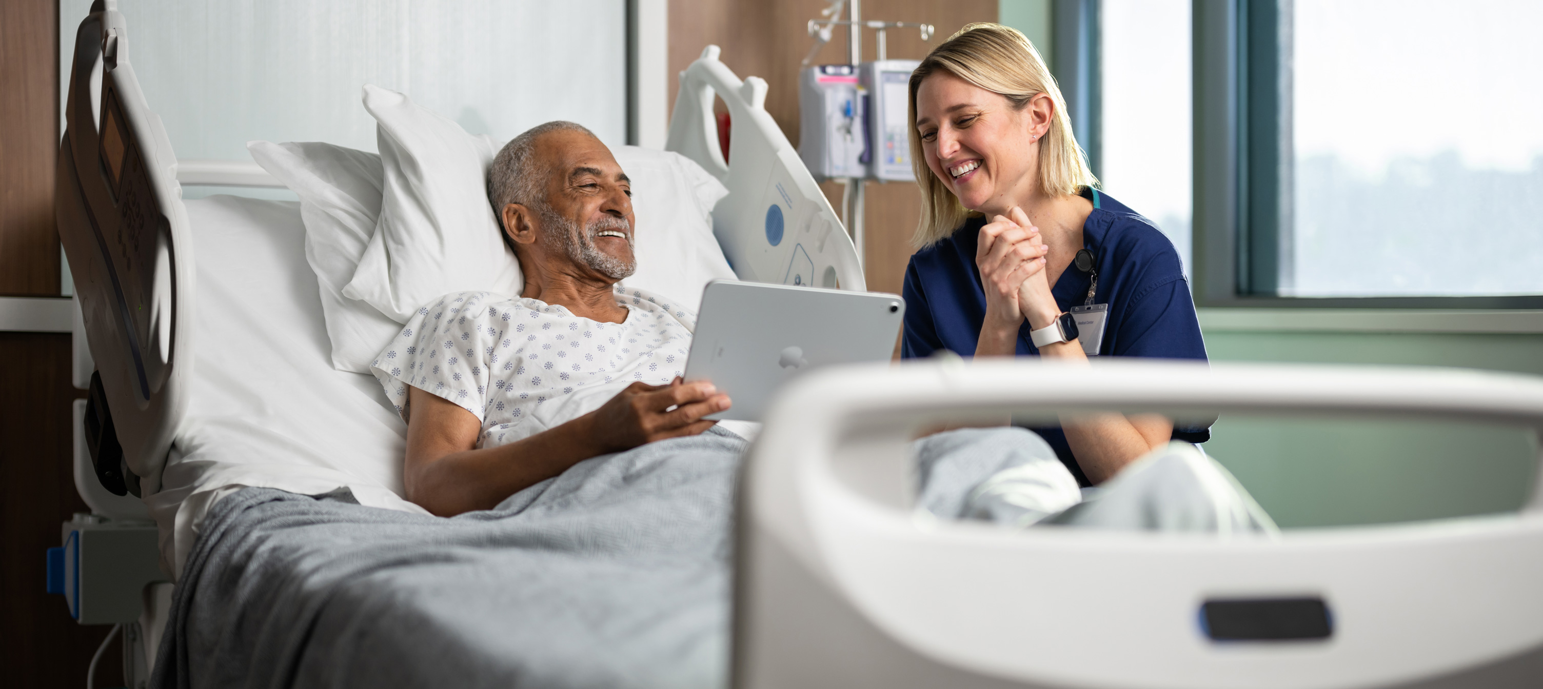 Ein Patient sitzt in einem Krankenhausbett. Er lächelt, während er mit einer Krankenpflegerin auf ein iPad Display schaut.